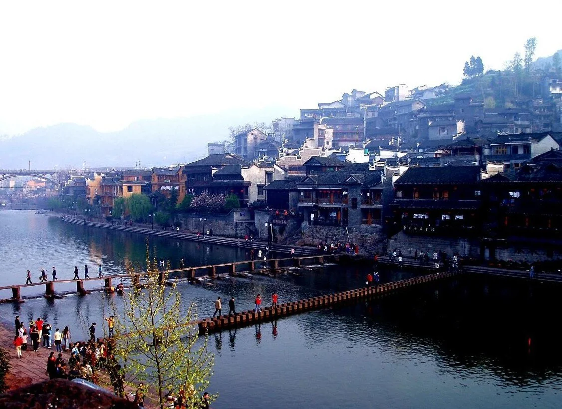 Visitors crossing stepping stones on the Tuojiang in Fenghuang—not Chadong—with riverfront buildings behind them.