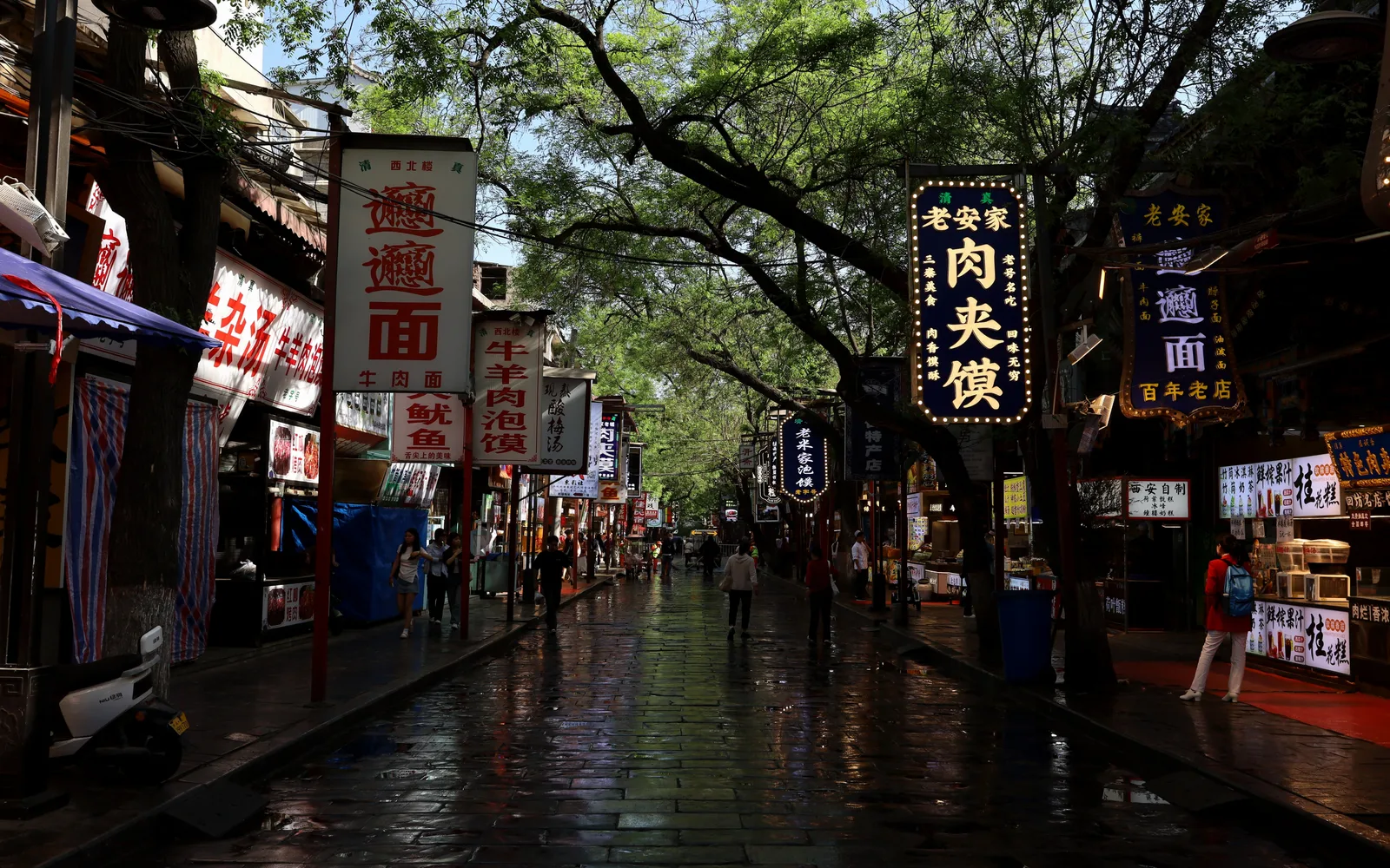 Rue pavée humide du quartier musulman de Xi’an, sous de grands arbres, bordée d’enseignes de nouilles au bœuf, roujiamo et soupe d’agneau.