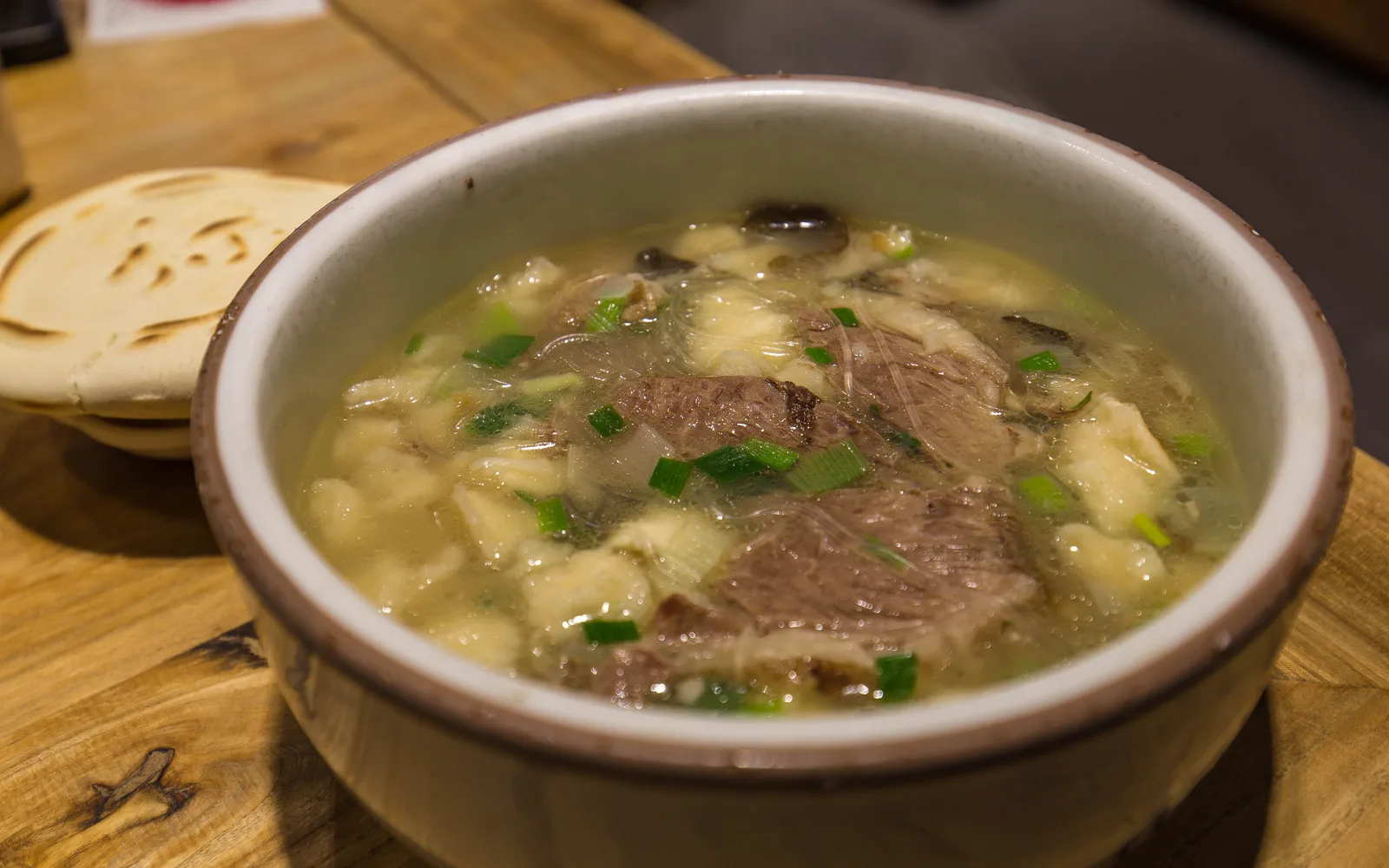 A bowl of yangrou paomo, lamb broth with pieces of flatbread and sliced lamb, beside a round flatbread on a wooden table