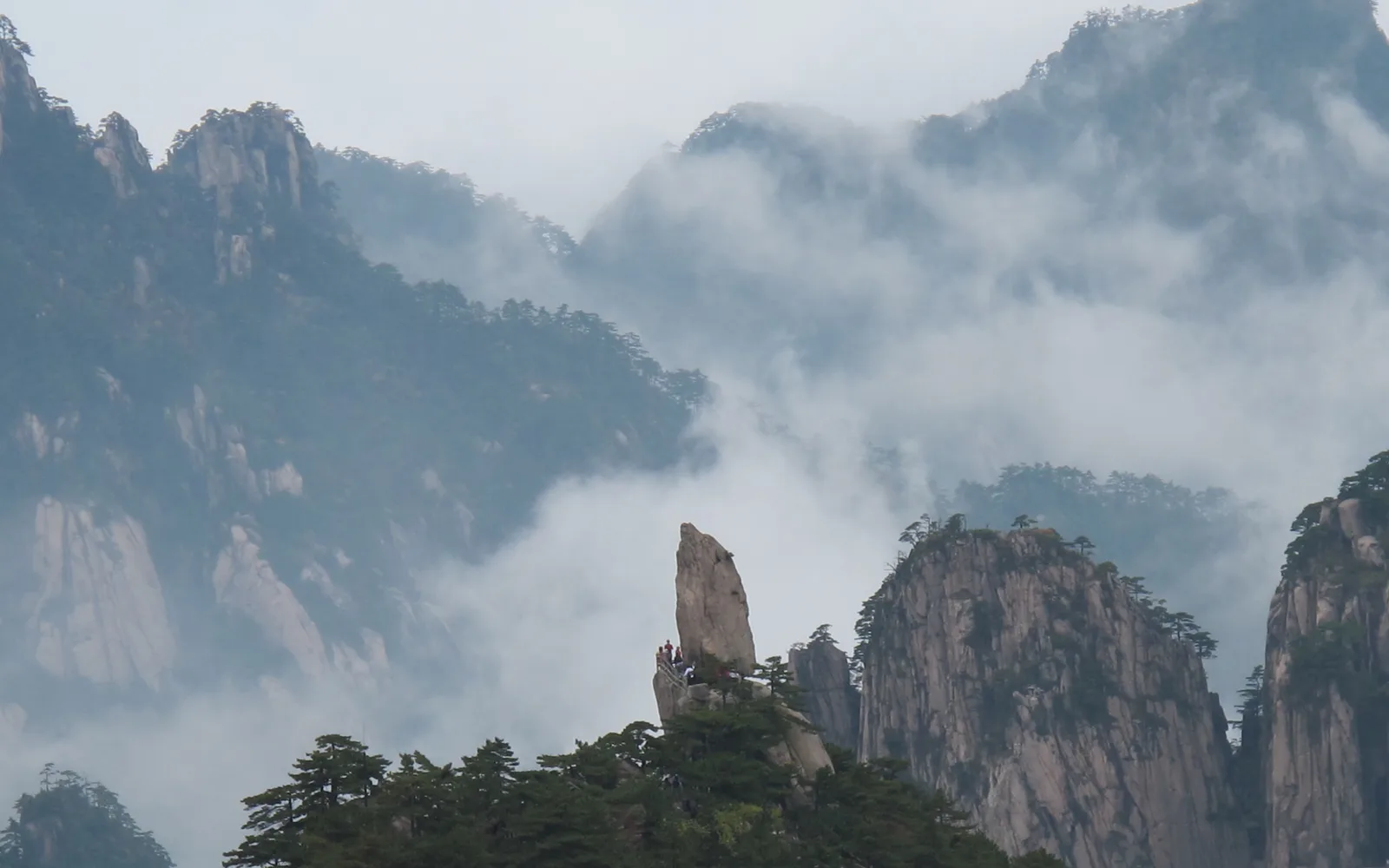 A tall standing rock on a Huangshan ridge with pines below it and cloud filling the valleys behind