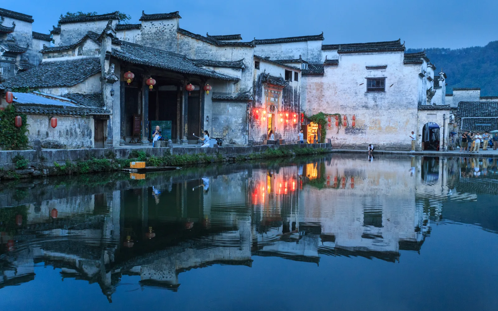White-walled Hui-style houses with red lanterns around Moon Pond in Hongcun at dusk, mirrored in the water