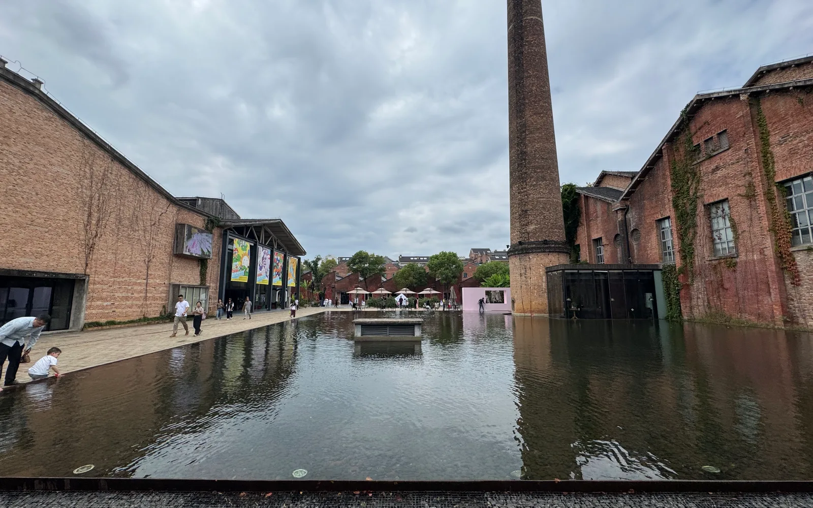 Brick factory halls and a tall chimney beside a reflecting pool at Taoxichuan in Jingdezhen, under a cloudy sky