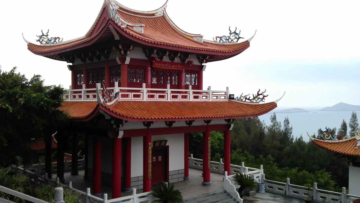 A red-and-white tower with an orange tiled roof at the Meizhou Mazu Ancestral Temple, with the sea in the distance.
