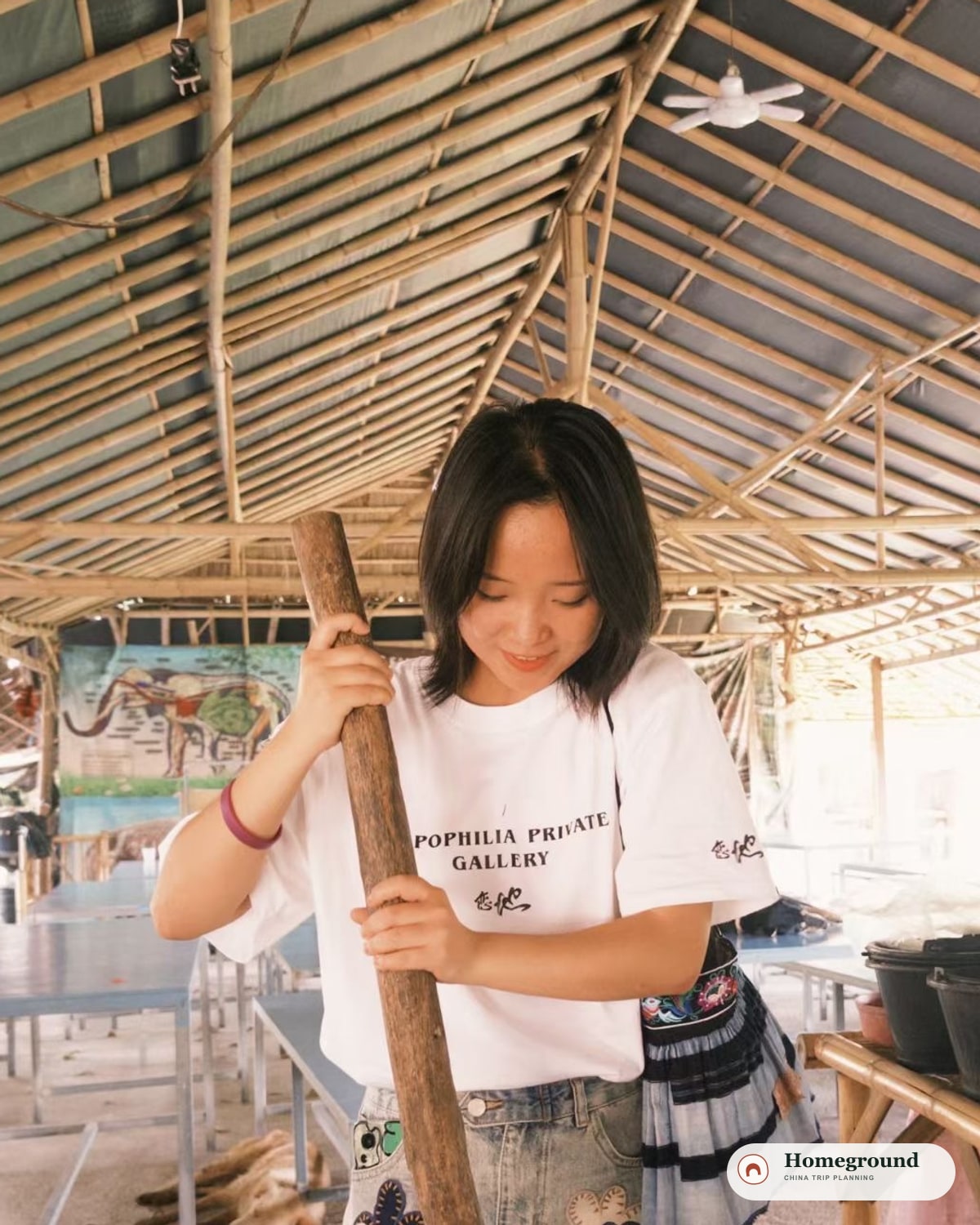 Tantan smiling while using a wooden pestle beneath a bamboo-roofed shelter.