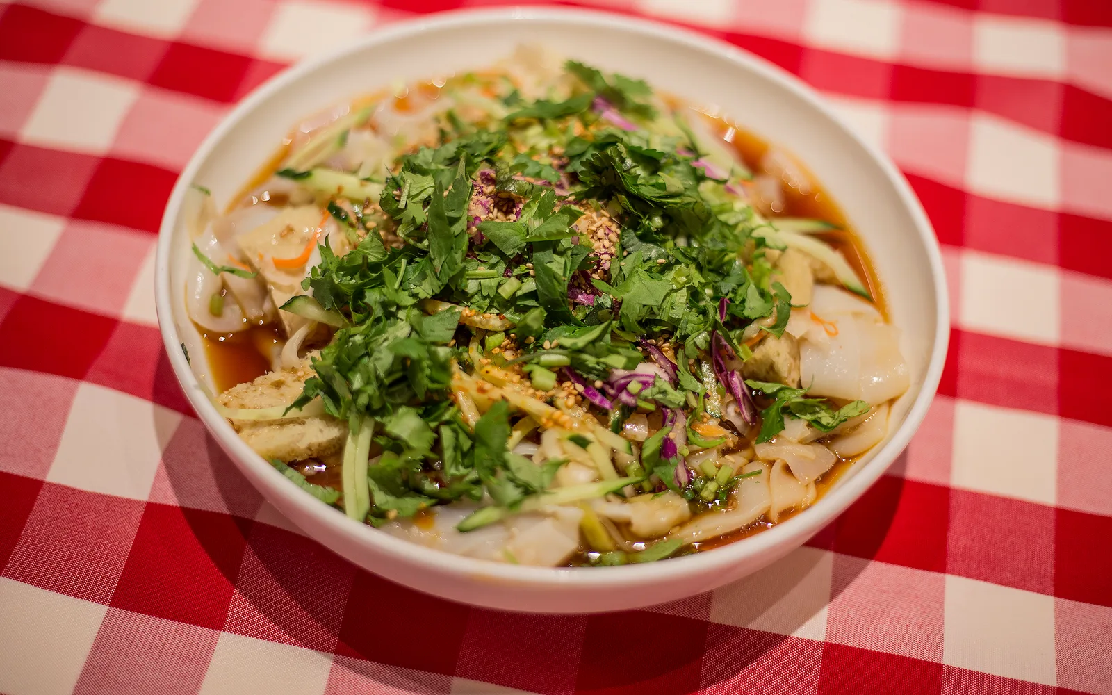 A bowl of liangpi cold noodles with coriander, cucumber and chilli oil on a red checked tablecloth