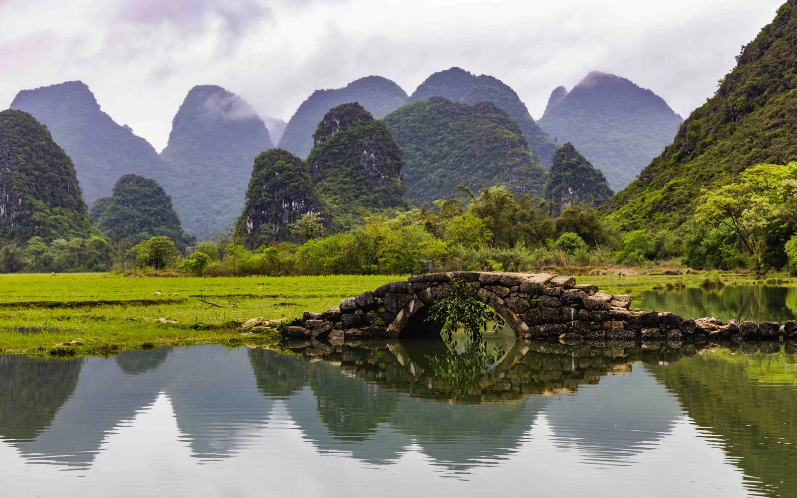 Vieux pont de pierre se reflétant dans l’eau, devant les reliefs karstiques de la campagne de Yangshuo.