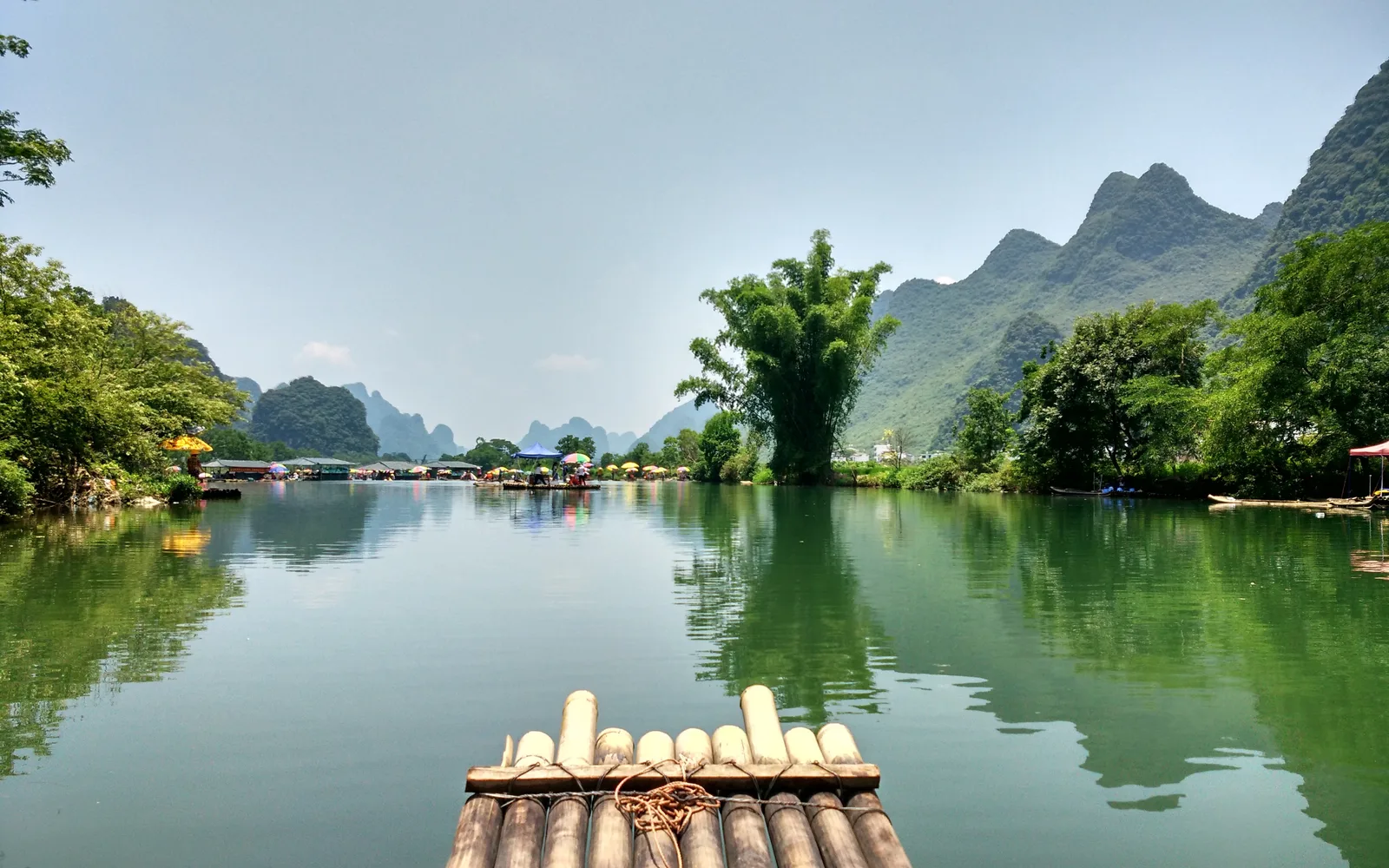 The bow of a bamboo raft on a calm green river in Yangshuo, with bamboo on the banks and karst peaks beyond